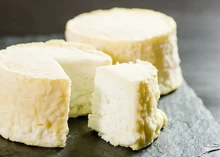 A partially cut wheel of Manchester cheese and a whole wheel, both showing a luscious cream line beneath a thin, wrinkly rind, rest on a black surface