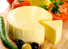A wheel of Kasseri cheese, partially sliced with a triangular piece removed, rests on a wooden board alongside green chilies, olives, and fresh tomatoes