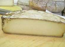 Half-cut wheel of Morlacco del Grappa cheese showcasing its thin rind and soft interior sits on wooden surface.
