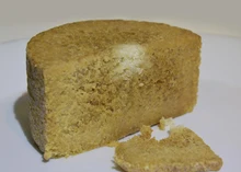 A large portion of Gammelost cheese highlighting its  golden-brown grainy texture, rests on a white surface