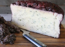 A wedge of Blu-61 cheese, showing blue veining and a layer of cranberries on top, placed alongside a cutting knife on a wooden surface