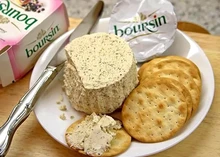 Boursin cheese spread on a cracker, with a large portion of Boursin cheese, on a white plate next to a box of the same cheese and a knife