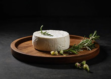 A disc of Goat cheese topped with rosemary herbs, sits on a circular wooden plate