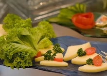 A disc and triangular pieces of Cream Havarti cheese arranged with lettuce leaves and tomato slices on a black plate, accompanied by a fork