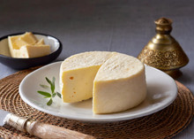 A wheel of cheese, partially sliced, sits on a white plate with a sprig of green garnish. A bowl with cheese pieces, a rustic knife, and a decorative container are also on the woven mat.