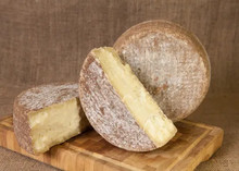 A whole wheel and half-cut wheels of Pecorino Ginepro cheese, showcasing their brown rind and pale white paste, are placed on a wooden board.