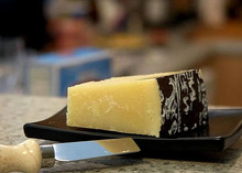 A black plate showcases a piece of Pecorino Romano cheese with its protective covering on one side, revealing its granular texture, accompanied by a knife