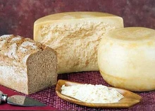 A whole wheel and a half-cut wheel of Reggianito cheese, along with grated Reggianito, crusty brown bread, and a cutting knife, are placed on a magenta mat.