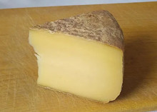 A wedge of Toma cheese with a rustic brown rind placed on a wooden board
