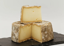 A large slice Tomme de Chevre placed on a partially cut wheel of it, showcasing its smooth interior and bluish-grey, bloomy rind, placed on black marble.