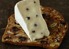 A piece of MouCo Truffello, revealing its velvety texture with black truffles, is placed on a dried fruit and nut cracker.