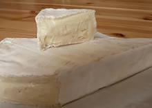 A large slice of Somerset Brie cheese, with a small piece placed on top of it revealing the white bloomy rind, rests on a wooden surface