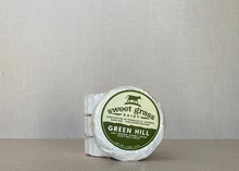 A round-shaped Green Hill cheese, wrapped in white paper with a label on top, placed next to three stacked Green Hill cheese wheels.