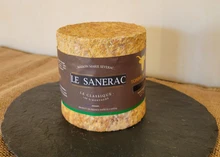 A cylindrical Le Sanerac cheese with a delicate whitish rind sits on a circular slab of black marble, all arranged atop a brown netted cloth.