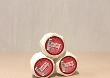 Three round-shaped Snow Camp cheeses, stacked and wrapped in white paper with a label on top, all placed on a beige surface.