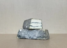 A slice of Sofia cheese placed on another slice of the same cheese, revealing its ash-colored exterior with a creamy interior, featuring two dark veins of vegetable ash streaking through