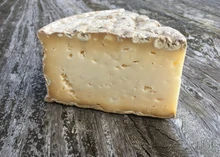 A triangular slice of Stoney Cross cheese, showcasing its creamy texture and mold-ripened rind, placed on a wooden surface.