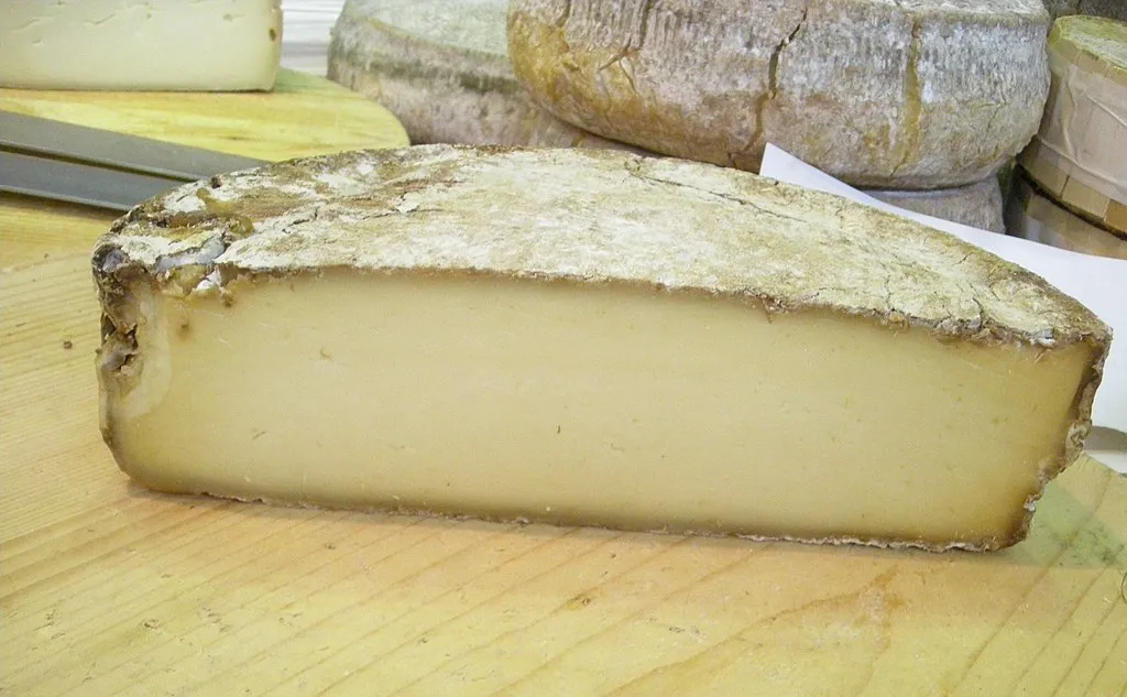 Half-cut wheel of Morlacco del Grappa cheese showcasing its thin rind and soft interior sits on wooden surface.
