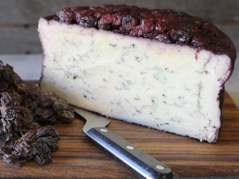 A wedge of Blu-61 cheese, showing blue veining and a layer of cranberries on top, placed alongside a cutting knife on a wooden surface