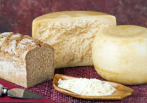 A whole wheel and a half-cut wheel of Reggianito cheese, along with grated Reggianito, crusty brown bread, and a cutting knife, are placed on a magenta mat.