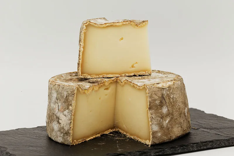 A large slice Tomme de Chevre placed on a partially cut wheel of it, showcasing its smooth interior and bluish-grey, bloomy rind, placed on black marble.
