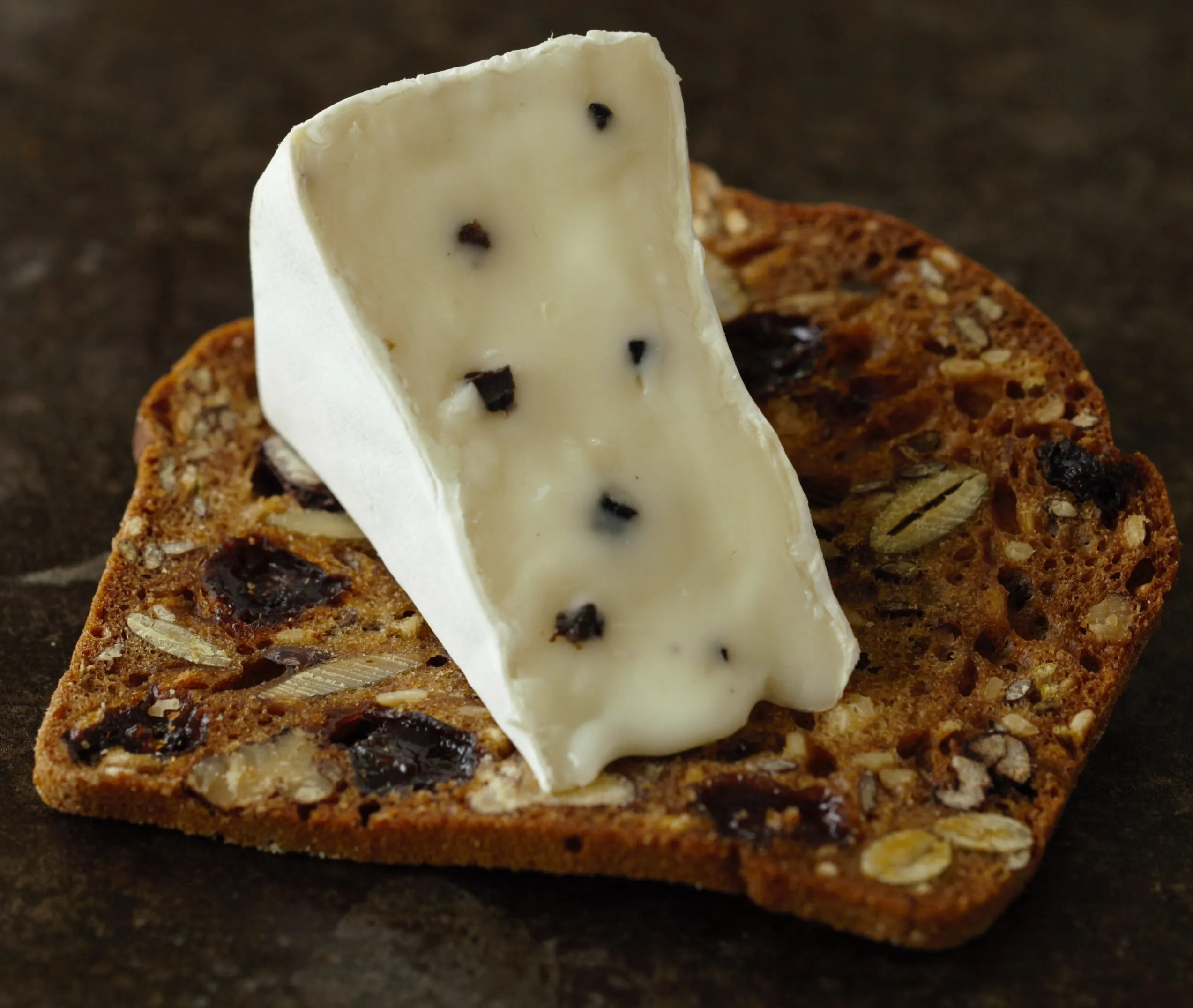 A piece of MouCo Truffello, revealing its velvety texture with black truffles, is placed on a dried fruit and nut cracker.