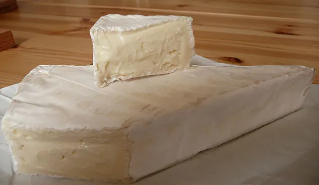 A large slice of Somerset Brie cheese, with a small piece placed on top of it revealing the white bloomy rind, rests on a wooden surface