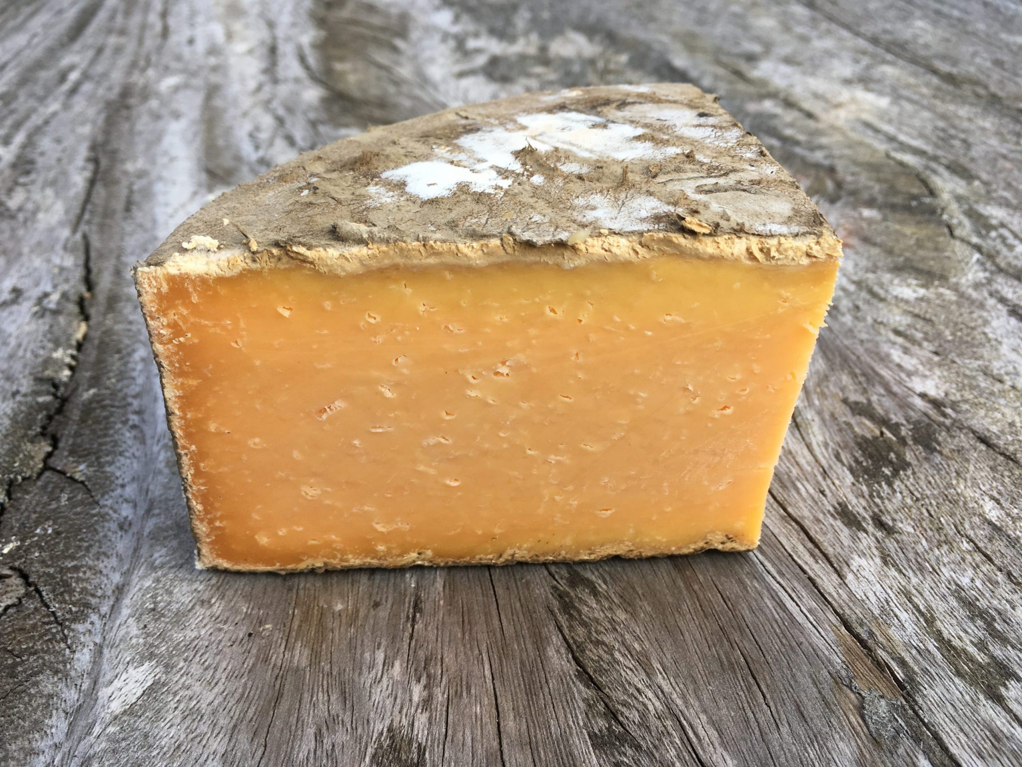 A slice of Double Gloucester cheese, showcasing its orange interior and natural rind, rests on a wooden surface
