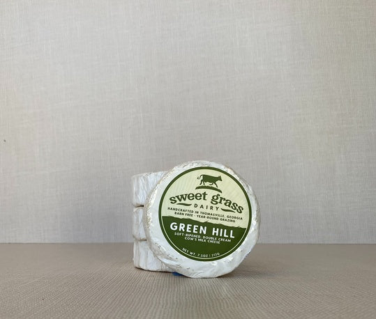 A round-shaped Green Hill cheese, wrapped in white paper with a label on top, placed next to three stacked Green Hill cheese wheels.