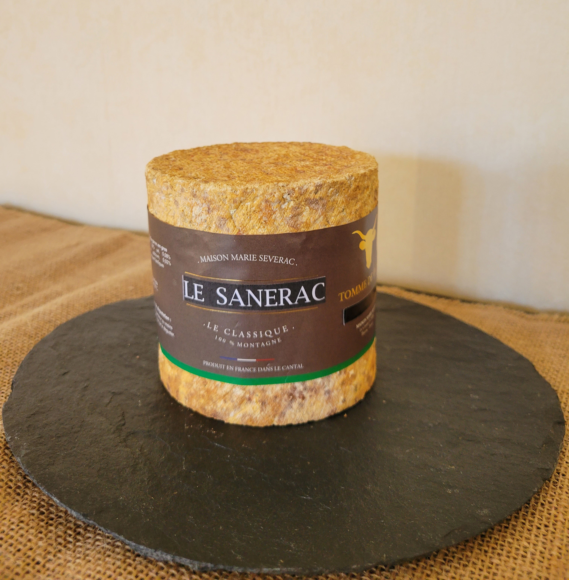 A cylindrical Le Sanerac cheese with a delicate whitish rind sits on a circular slab of black marble, all arranged atop a brown netted cloth.