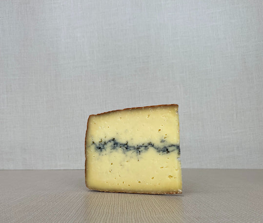 A slice of Ridgeline cheese, revealing its dark rind and pale yellow interior with a dark vein of vegetable ash streaking through the middle