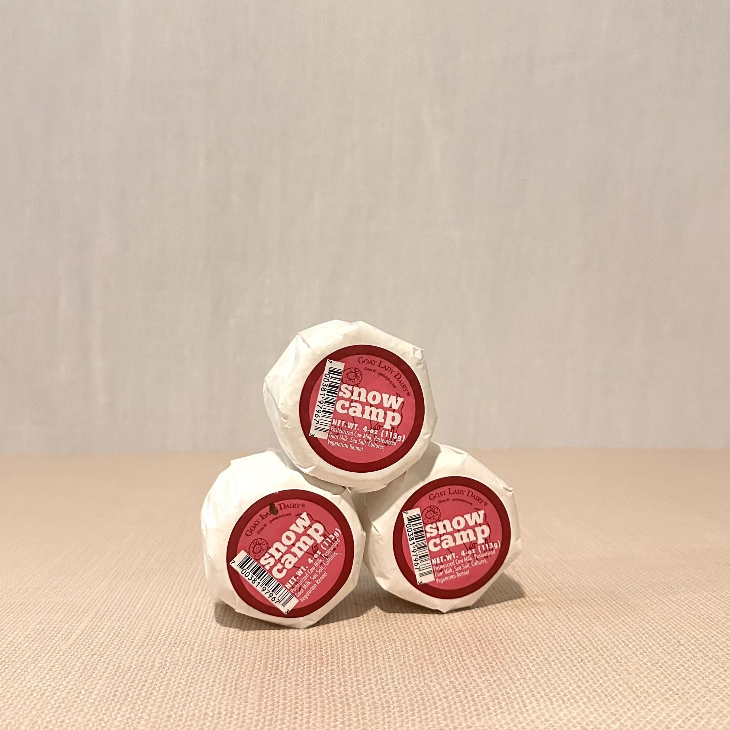 Three round-shaped Snow Camp cheeses, stacked and wrapped in white paper with a label on top, all placed on a beige surface.