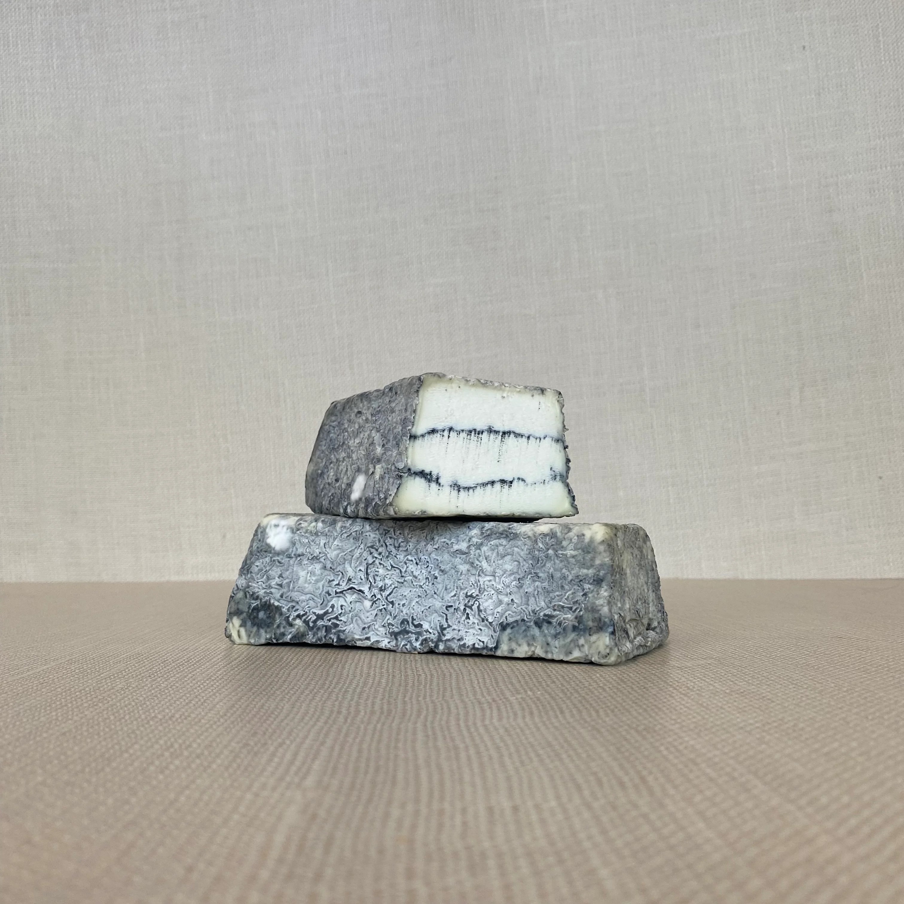 A slice of Sofia cheese placed on another slice of the same cheese, revealing its ash-colored exterior with a creamy interior, featuring two dark veins of vegetable ash streaking through
