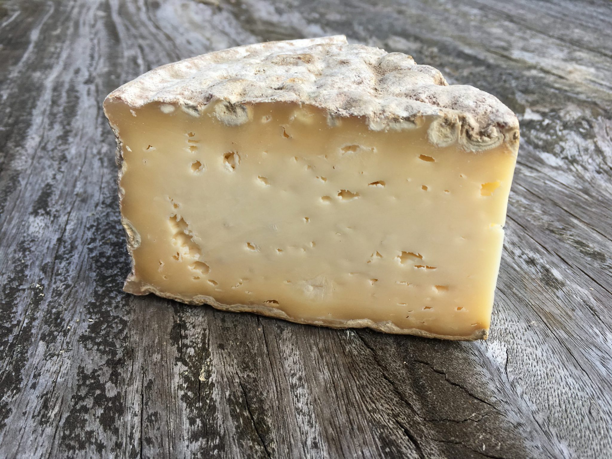 A triangular slice of Stoney Cross cheese, showcasing its creamy texture and mold-ripened rind, placed on a wooden surface.